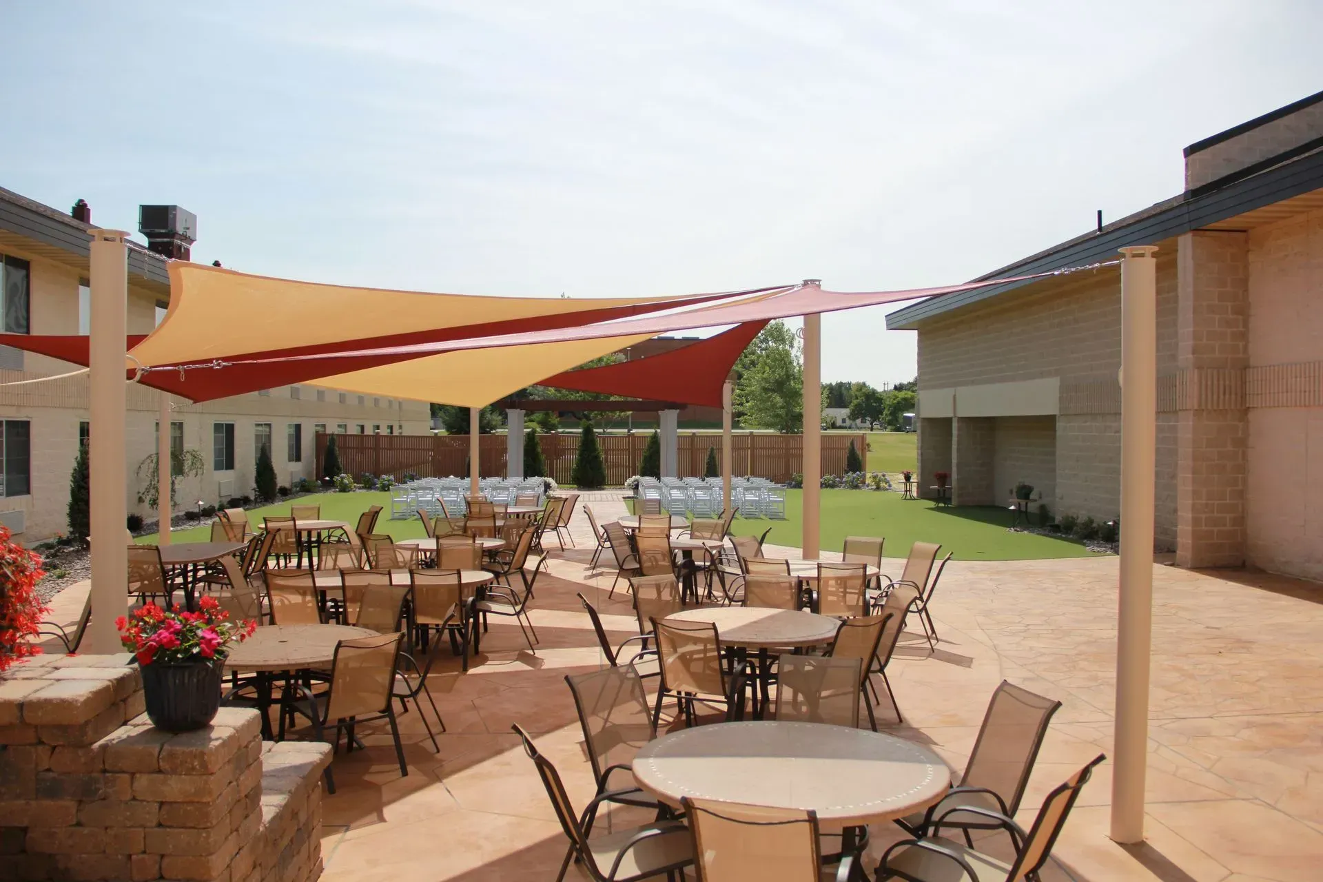 Outdoor patio with tables, chairs, and shade sails; lawn and building in background.
