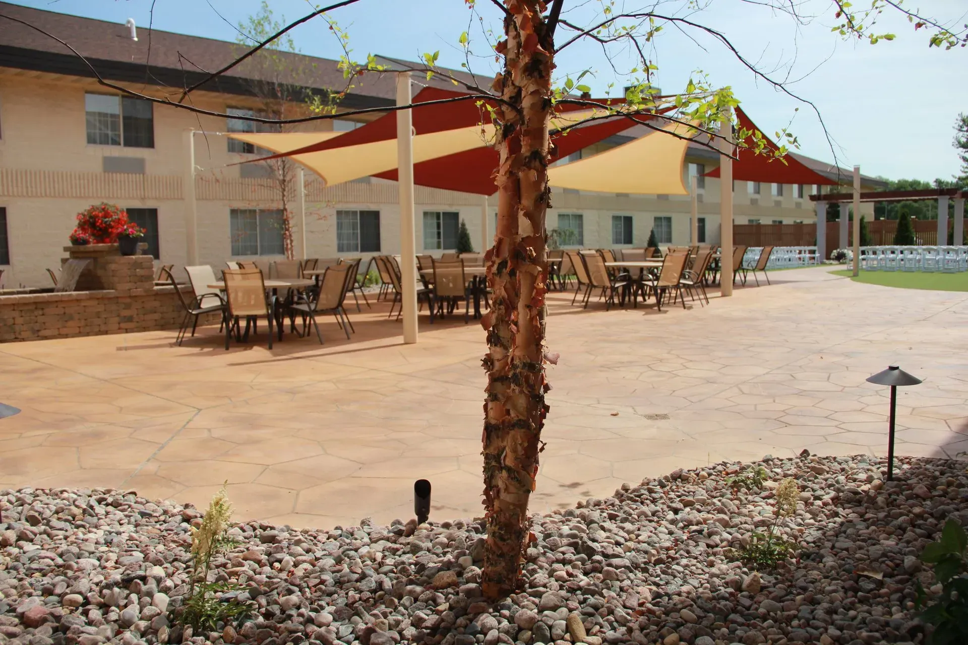 Outdoor patio with tables, chairs, and shade sails. Building in the background, rock garden in the foreground.