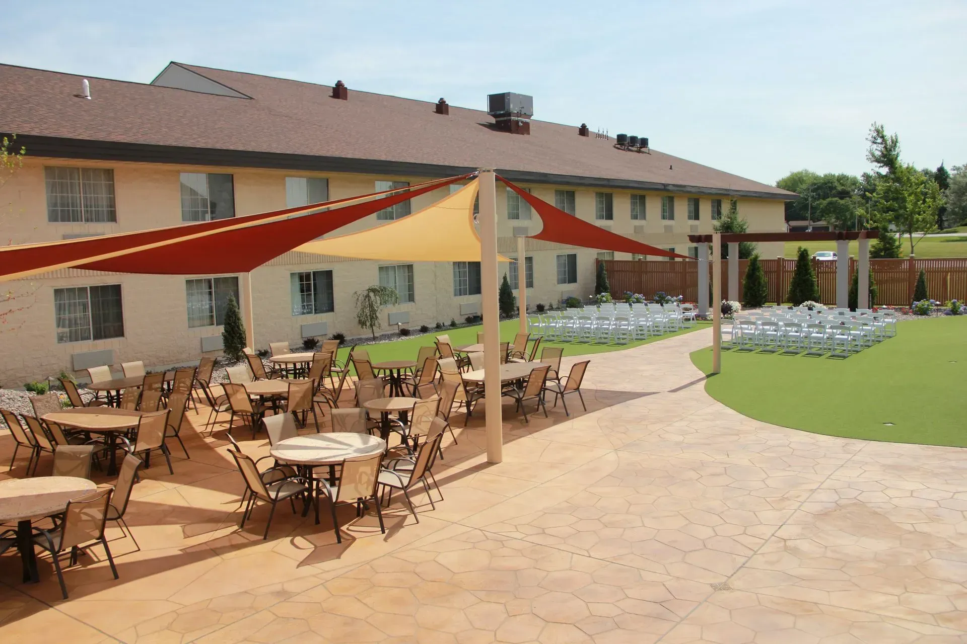 Outdoor dining area with tables, chairs, and shade sails in front of a light-colored building.