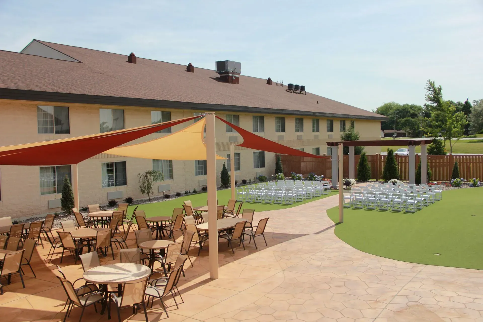 Outdoor event space with tables, chairs, and shade sails, adjacent to a two-story building.