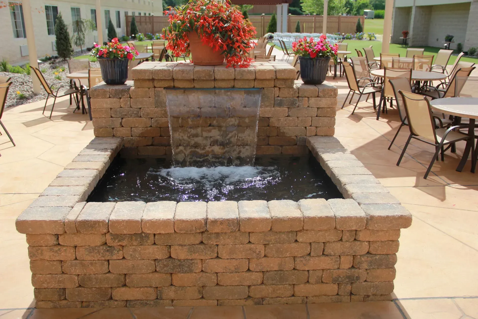 Water fountain built with tan bricks, with flowers, and tables and chairs in the background.