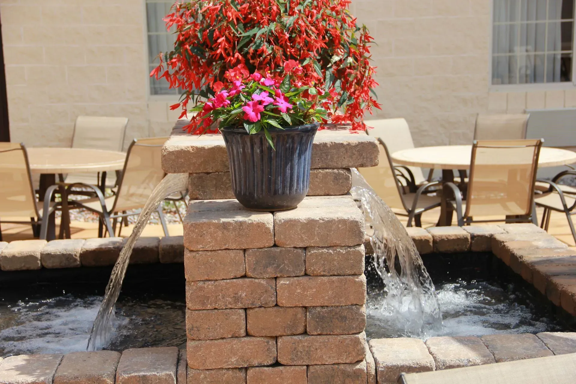 A tiered brick fountain with a potted plant, water flowing, patio furniture in the background.