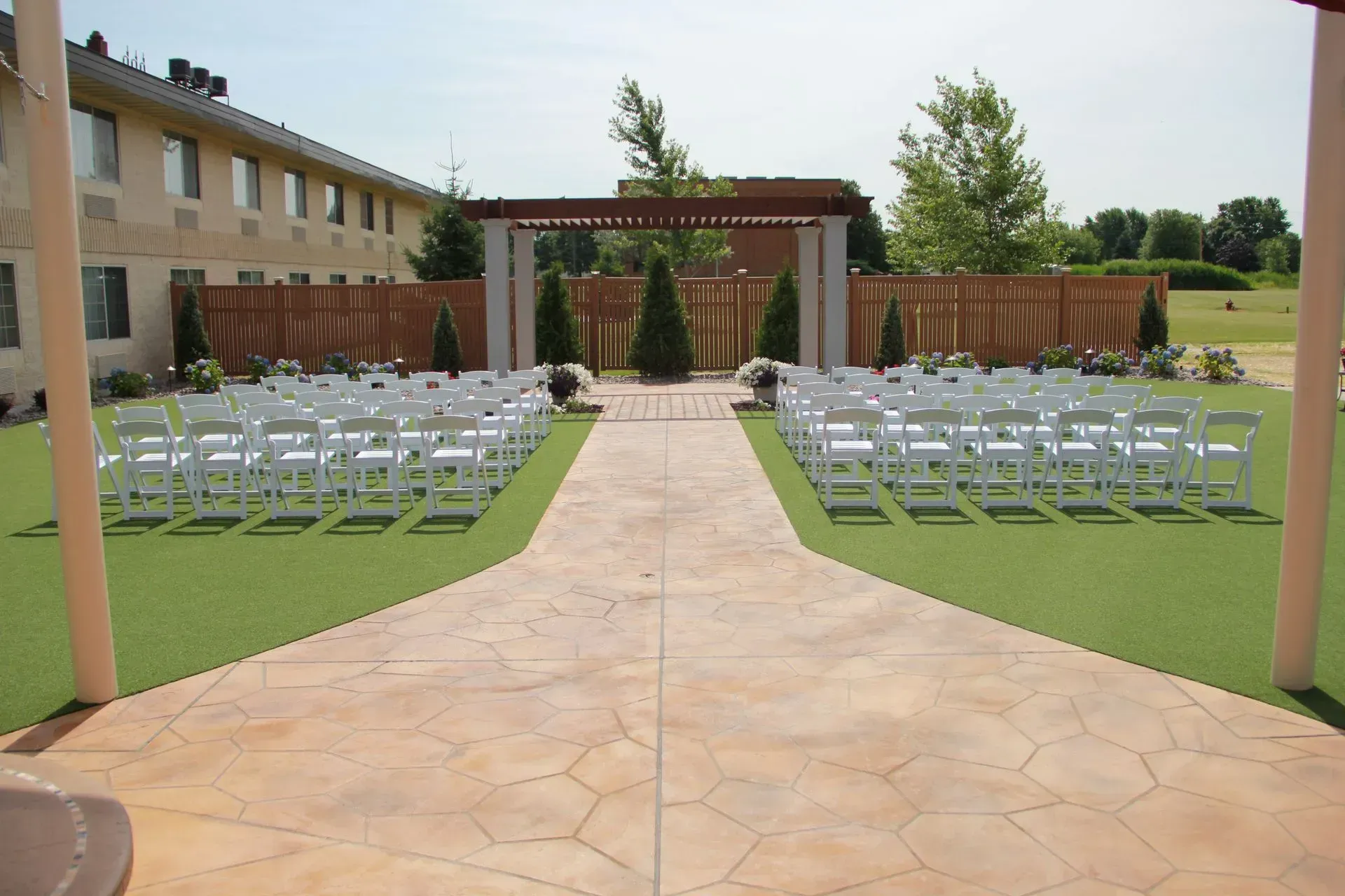 Outdoor wedding ceremony setup with white chairs on green turf, leading to a wooden arbor.