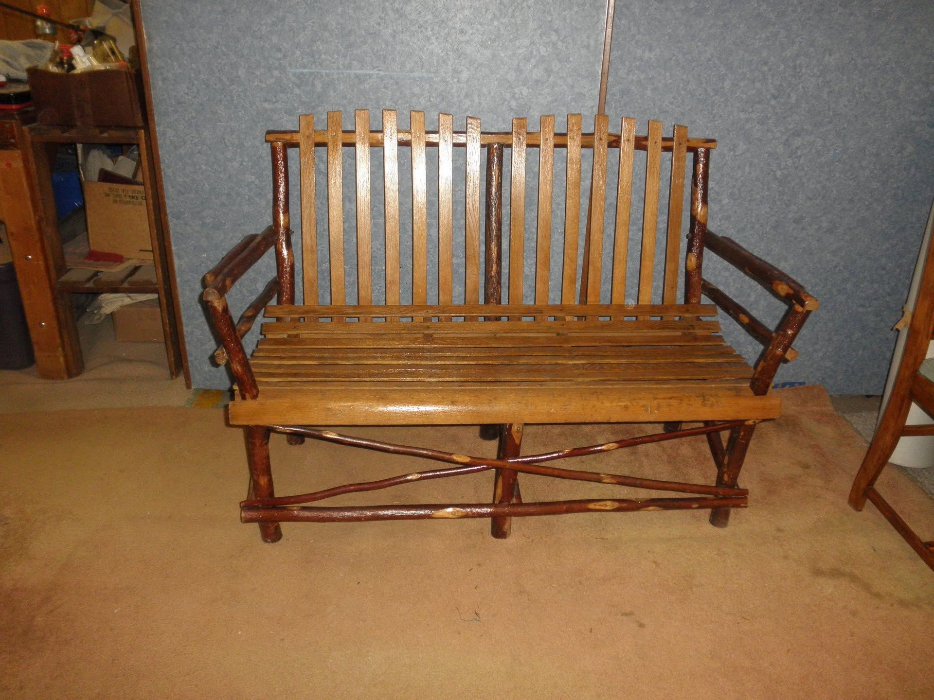 Wooden slat bench with armrests and cross-braced base on a tan floor against a gray wall.