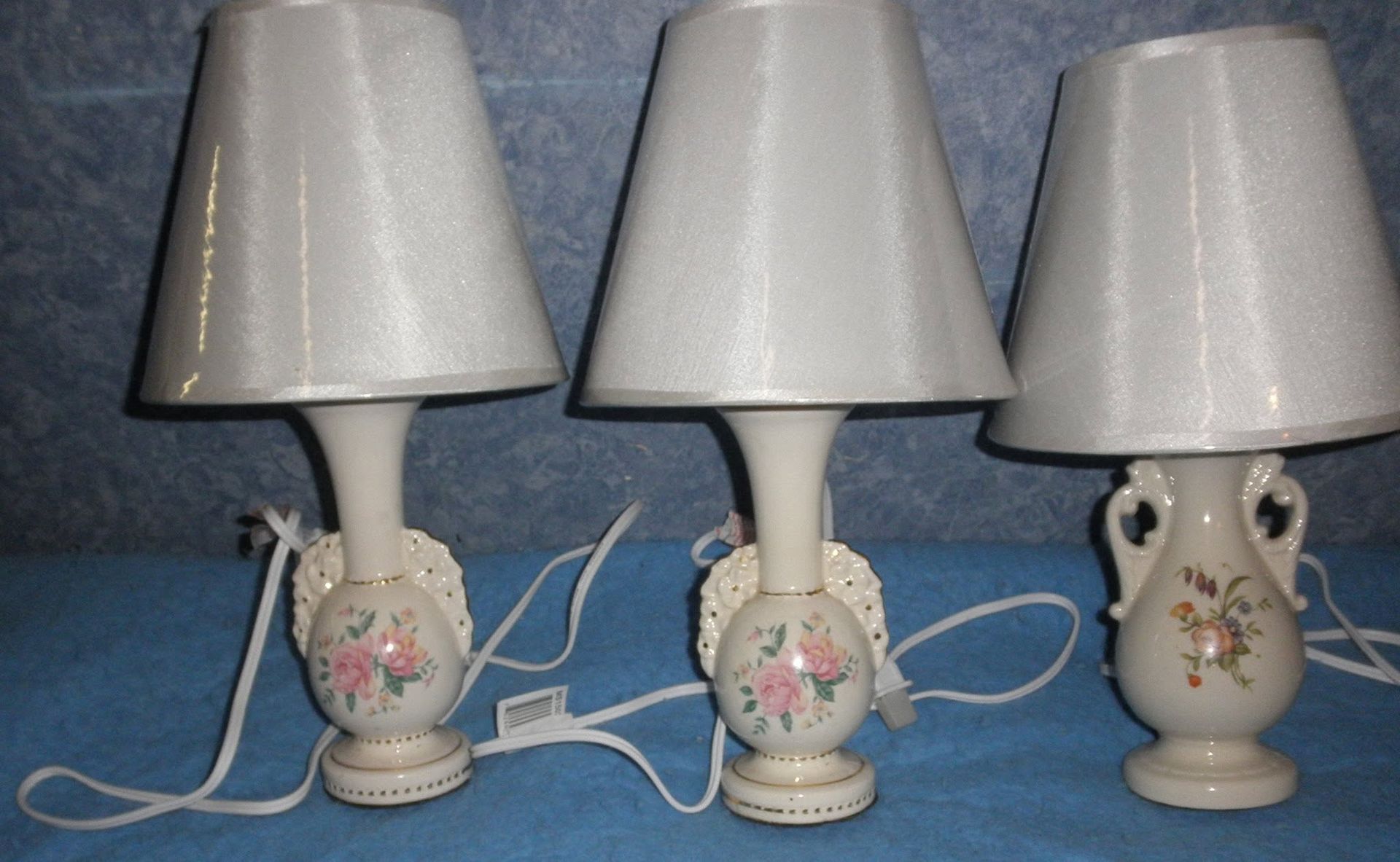 Three small ceramic table lamps with white shades and floral bases lined up on a blue surface.