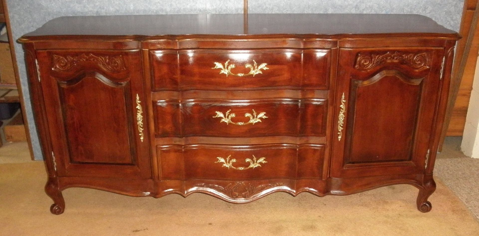 Ornate dark wood dresser with three drawers and two cabinet doors against a brick wall