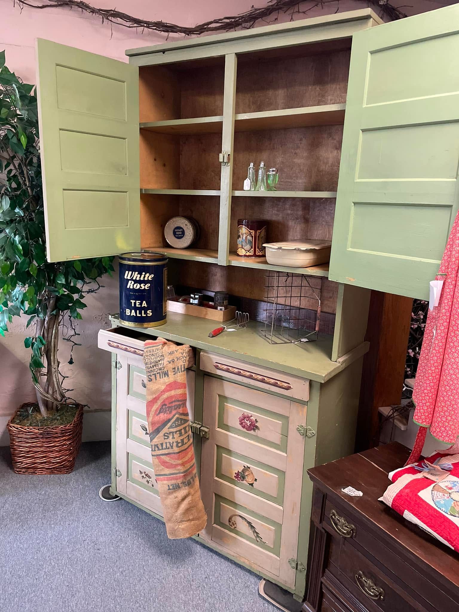 Open mint-green kitchen cabinet with shelves, drawers, and dishes in a cozy room
