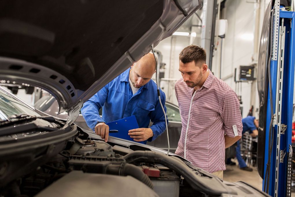 Two mechanics in a garage inspect an open car engine, one holding a blue tablet.