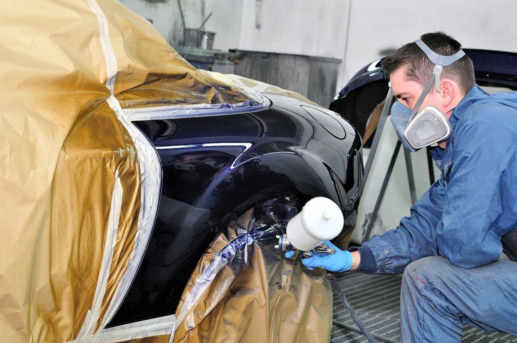 Worker spray-painting a black car in an auto body shop, masked and wearing gloves
