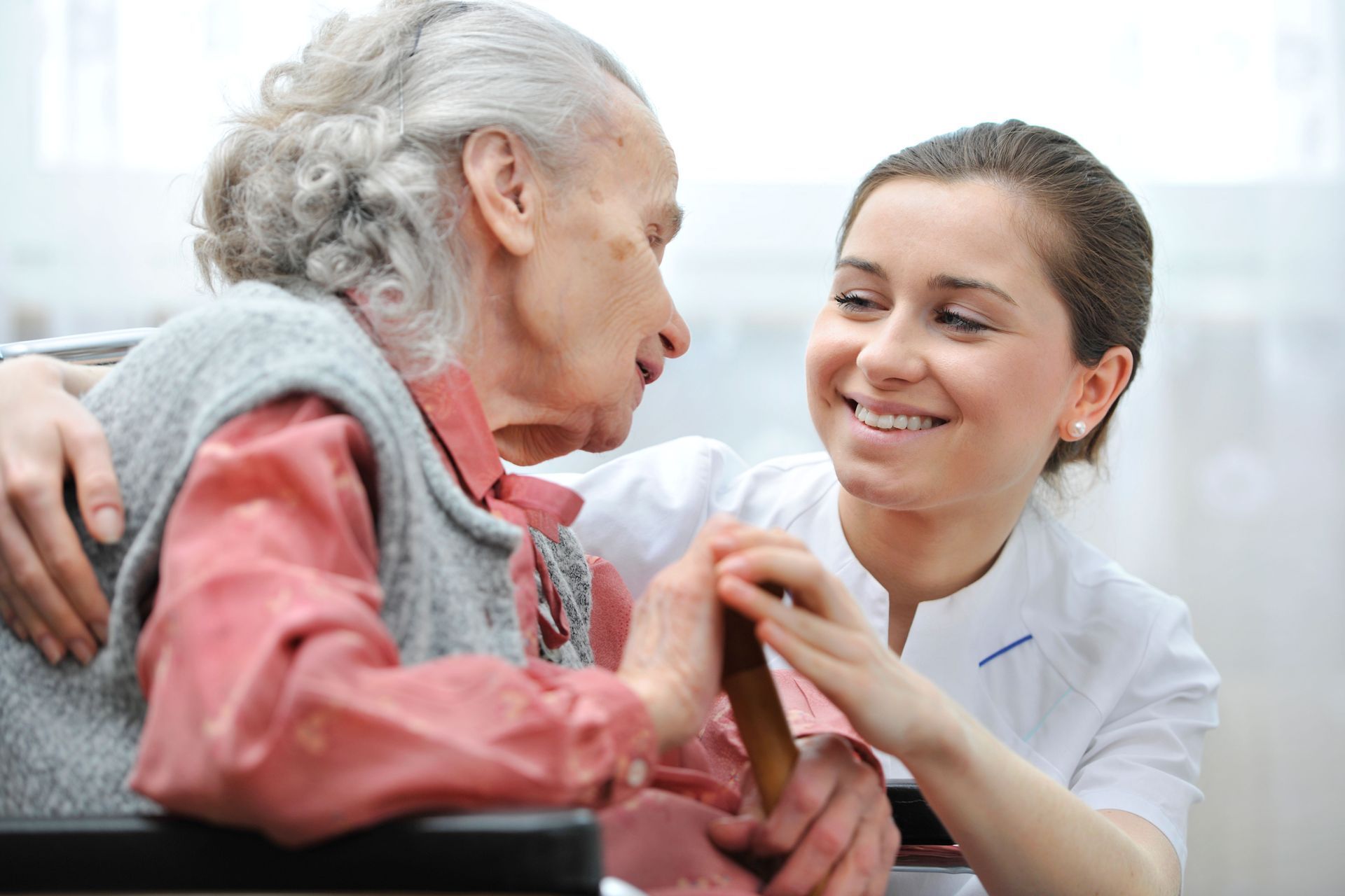A care provider smiling while holding hands with and offering support to a person in a wheelchair.