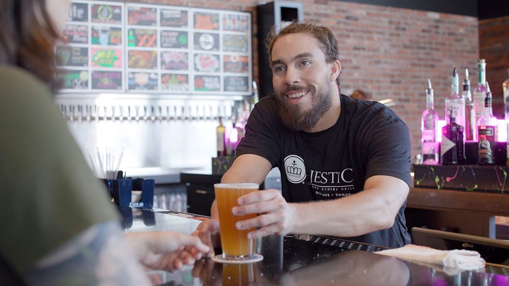 A bartender with a beard smiles, handing a beer to a customer at a bar.