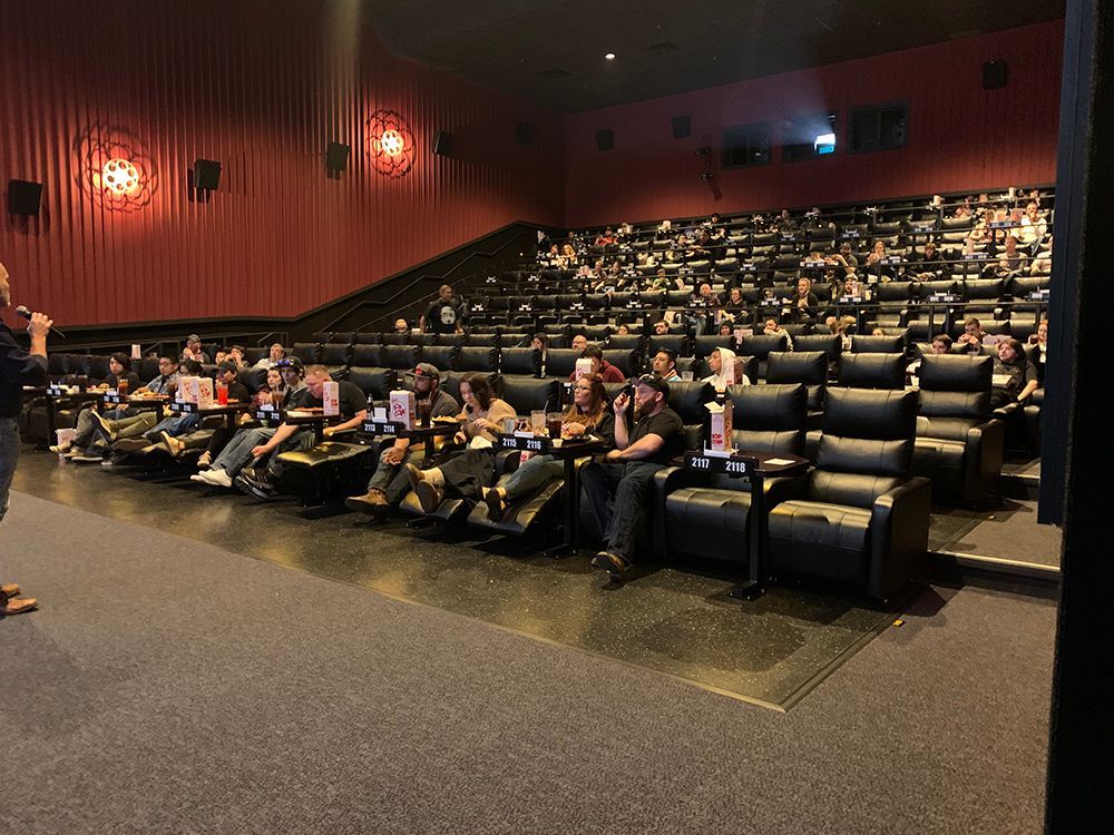 Movie theater with audience seated, watching a presentation from a person on stage. Red walls, black seats.