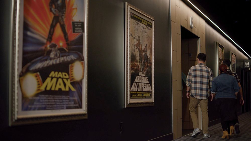 Two people walk past movie posters in a theater hallway;