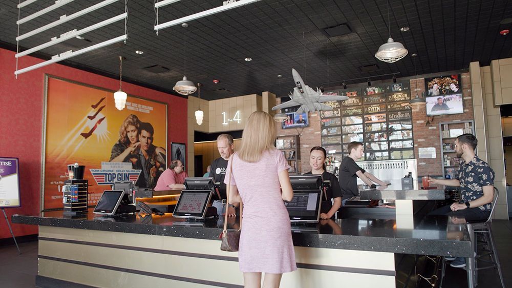 A woman in a pink dress orders at a movie theater counter. Posters for films and a model plane are visible.