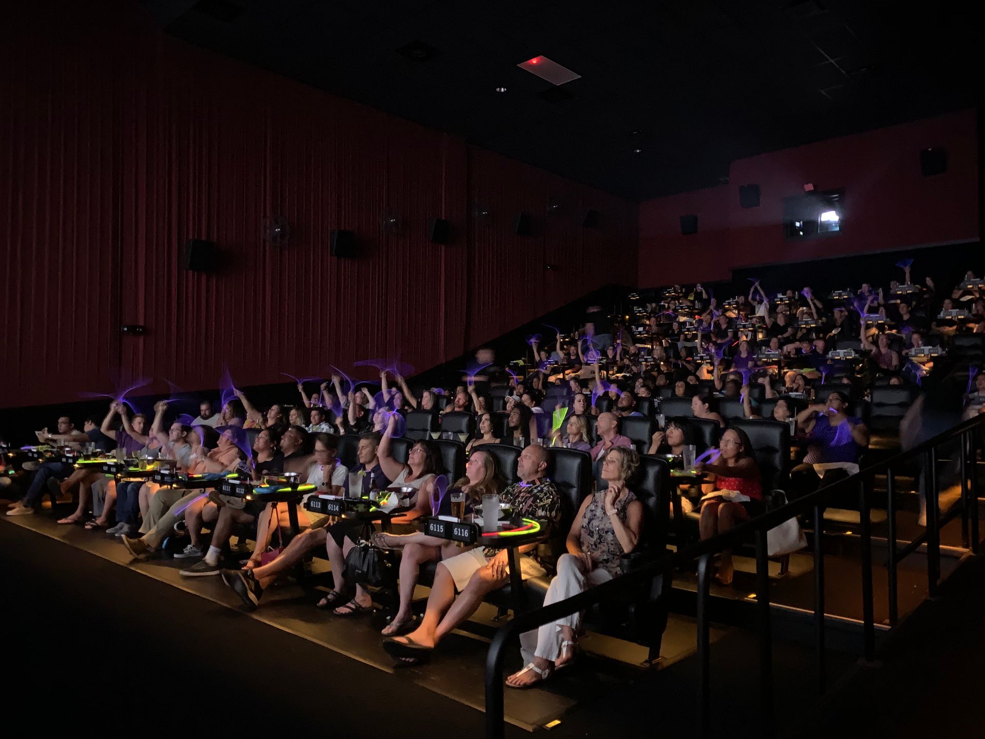 A crowded movie theater. Audience members wave purple objects; red curtain background.