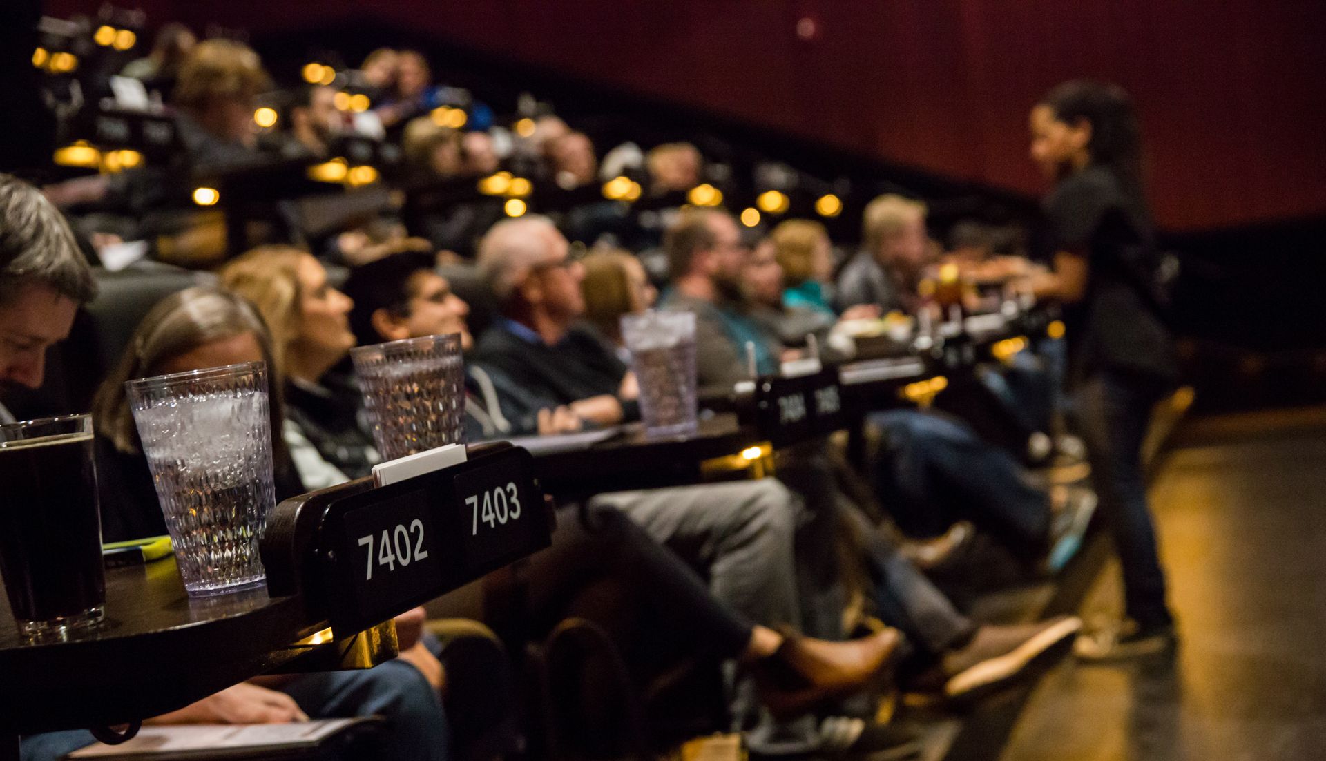People watching a movie in a theater with servers. Some are eating or drinking. Low lighting.