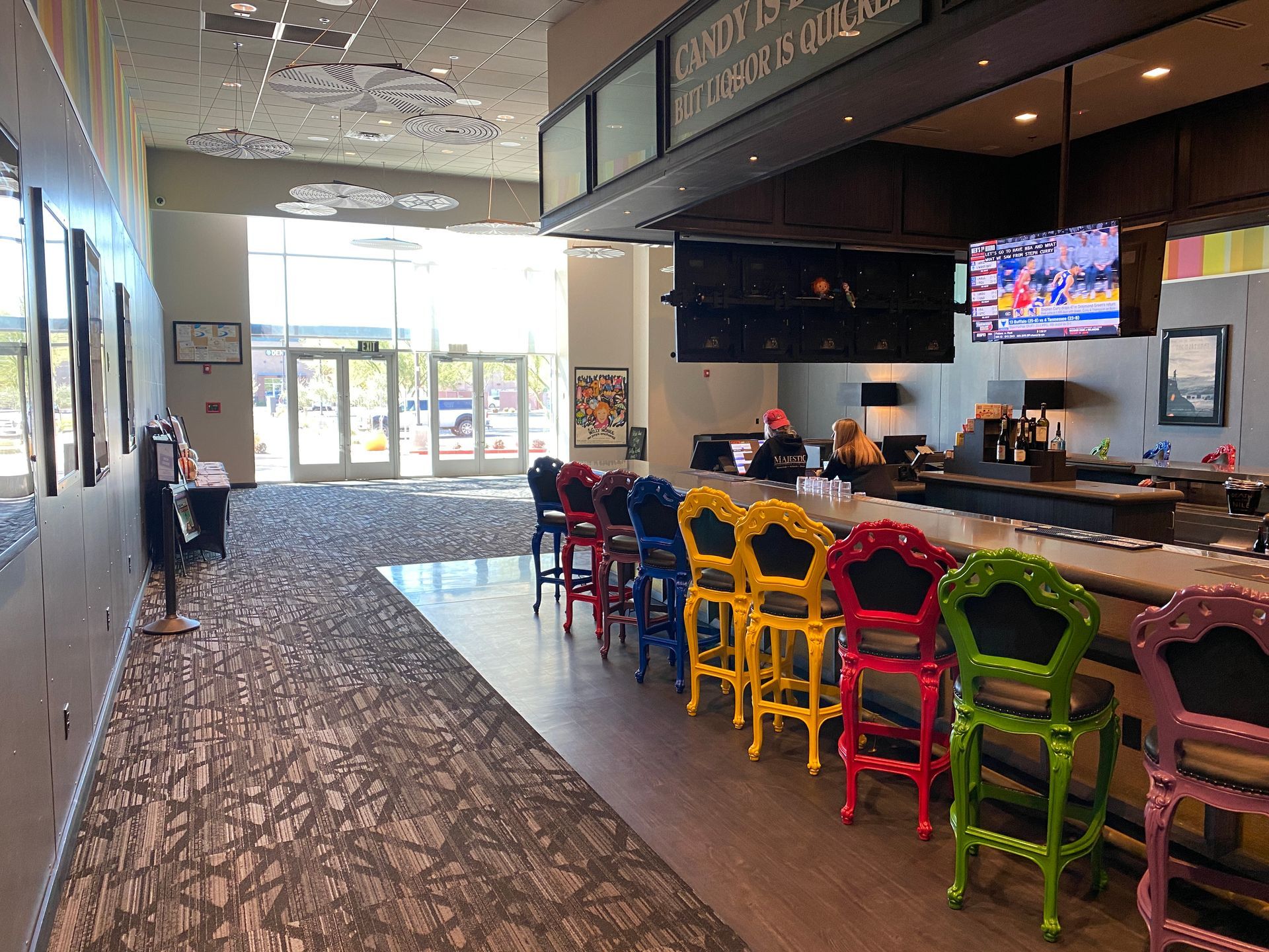 Bar area with colorful chairs, bar with customers, and doors leading outside.