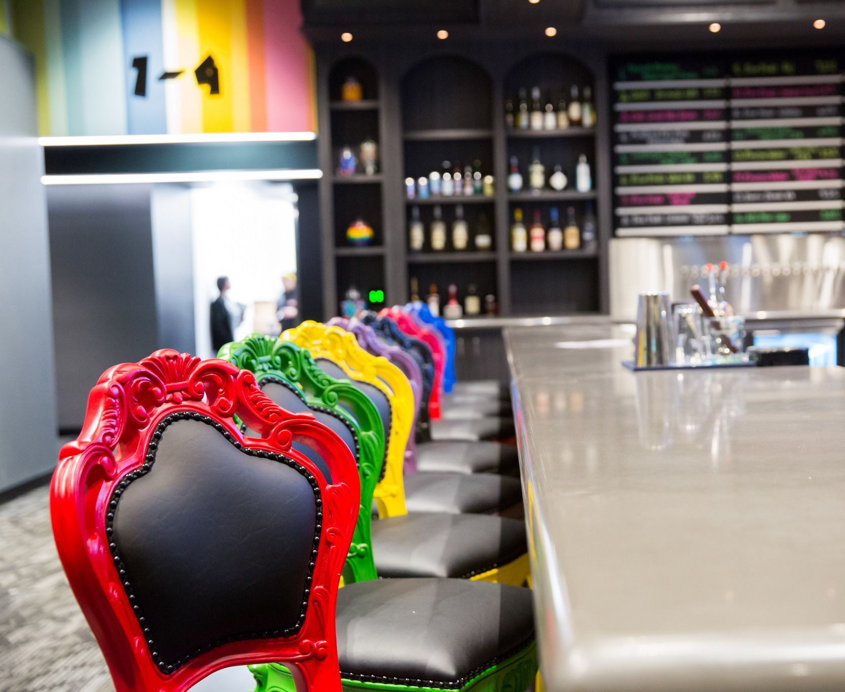 Colorful bar stools line a bar with a gray countertop and a shelf of bottles against a dark wall.