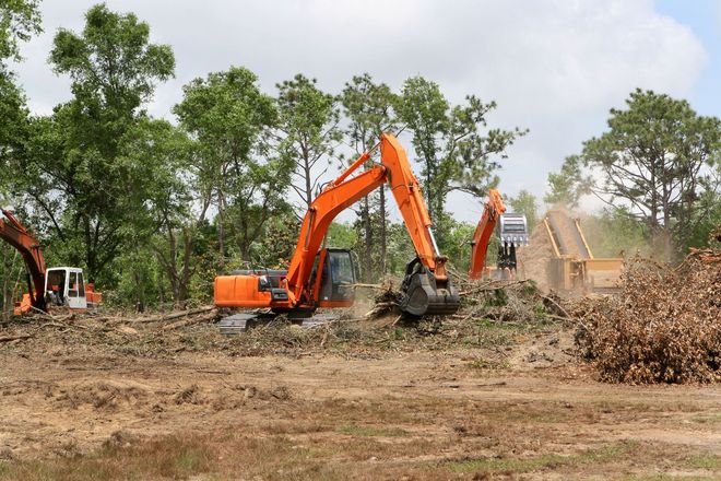 Orange construction excavators clear trees and vegetation from a muddy field under a partly cloudy sky.