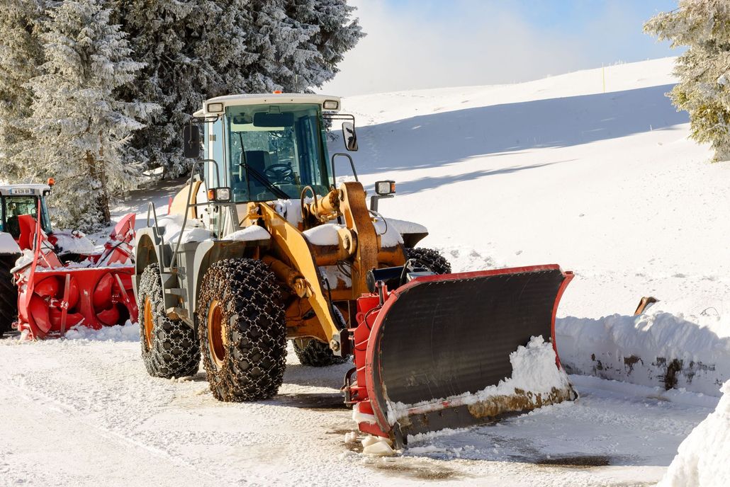A yellow tractor with a large plow and tire chains parked on a snowy road beside a red snowblower, with trees in the back.