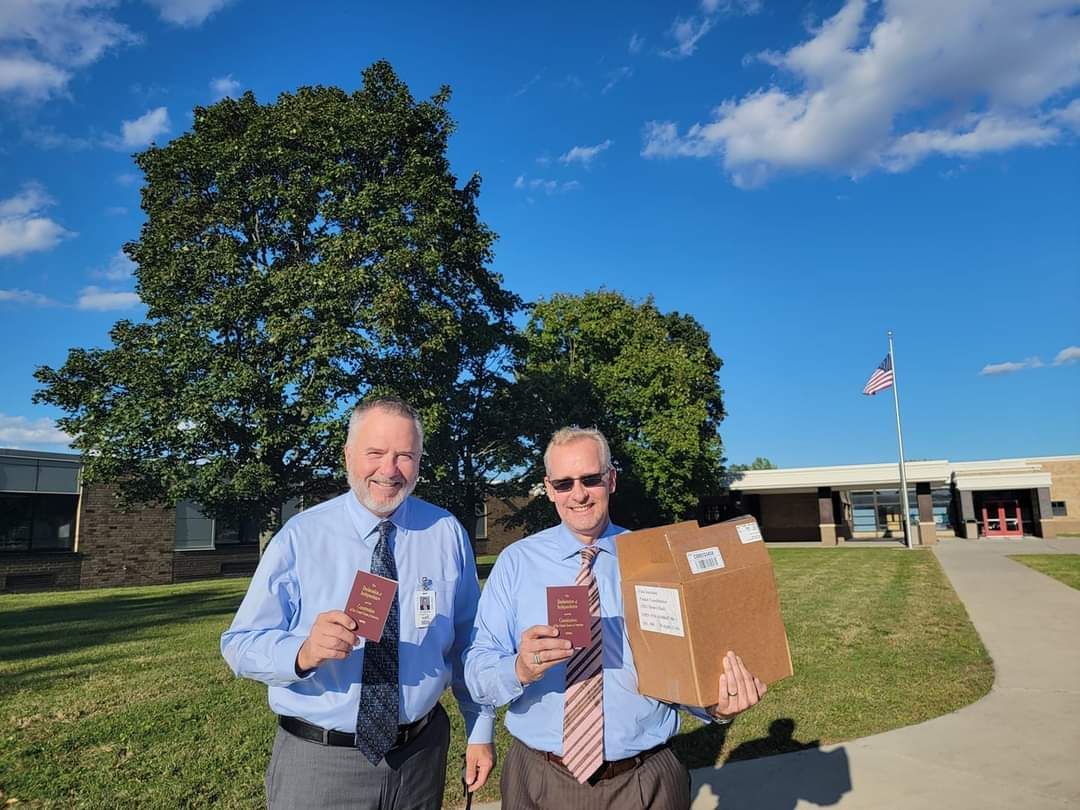 Two men are standing next to each other in front of a school holding a cardboard box.