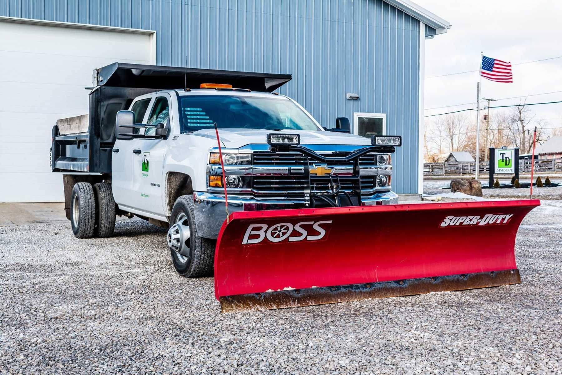 White dump truck with red snow plow parked beside a metal building in a gravel lot