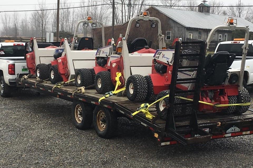 Several red and white skid-steer loaders strapped on a trailer in a gravel lot, with one operator standing beside them