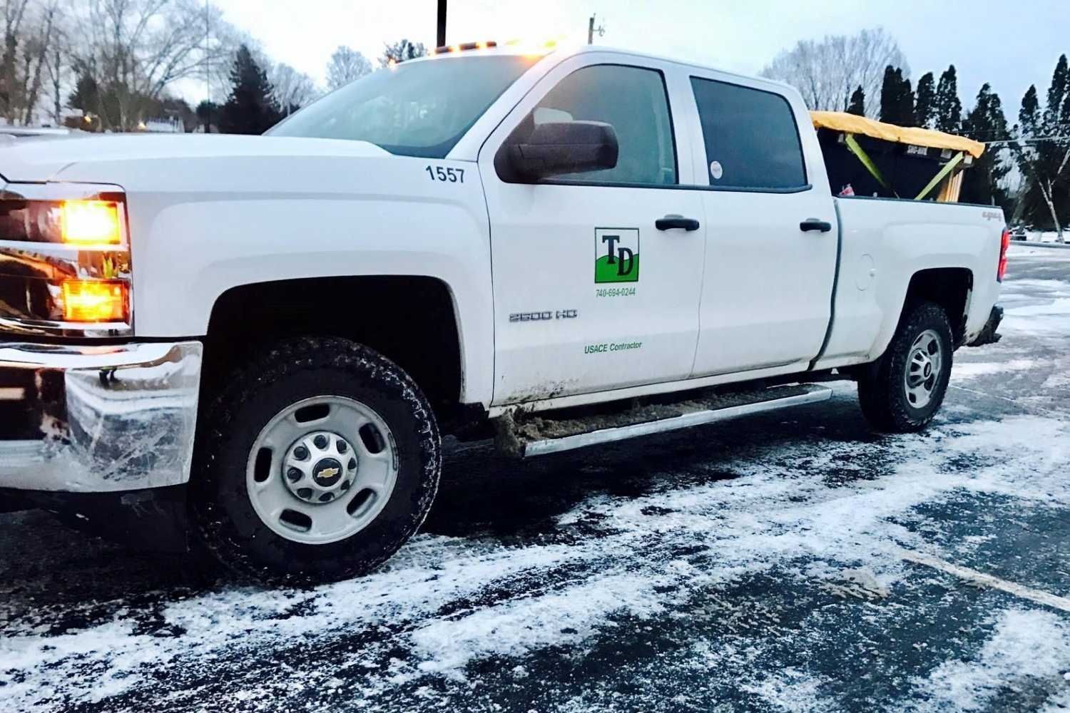 White pickup truck parked on a snowy lot, with a green logo on the door and yellow equipment in the bed