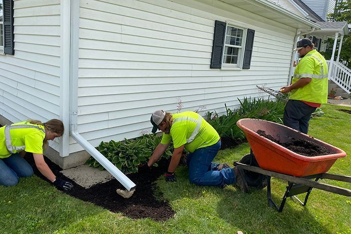 Three workers in neon shirts dig a trench beside a house; one pushes a wheelbarrow.