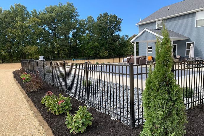 Black metal fence around gravel patio beside a gray house, with landscaped shrubs and trees.