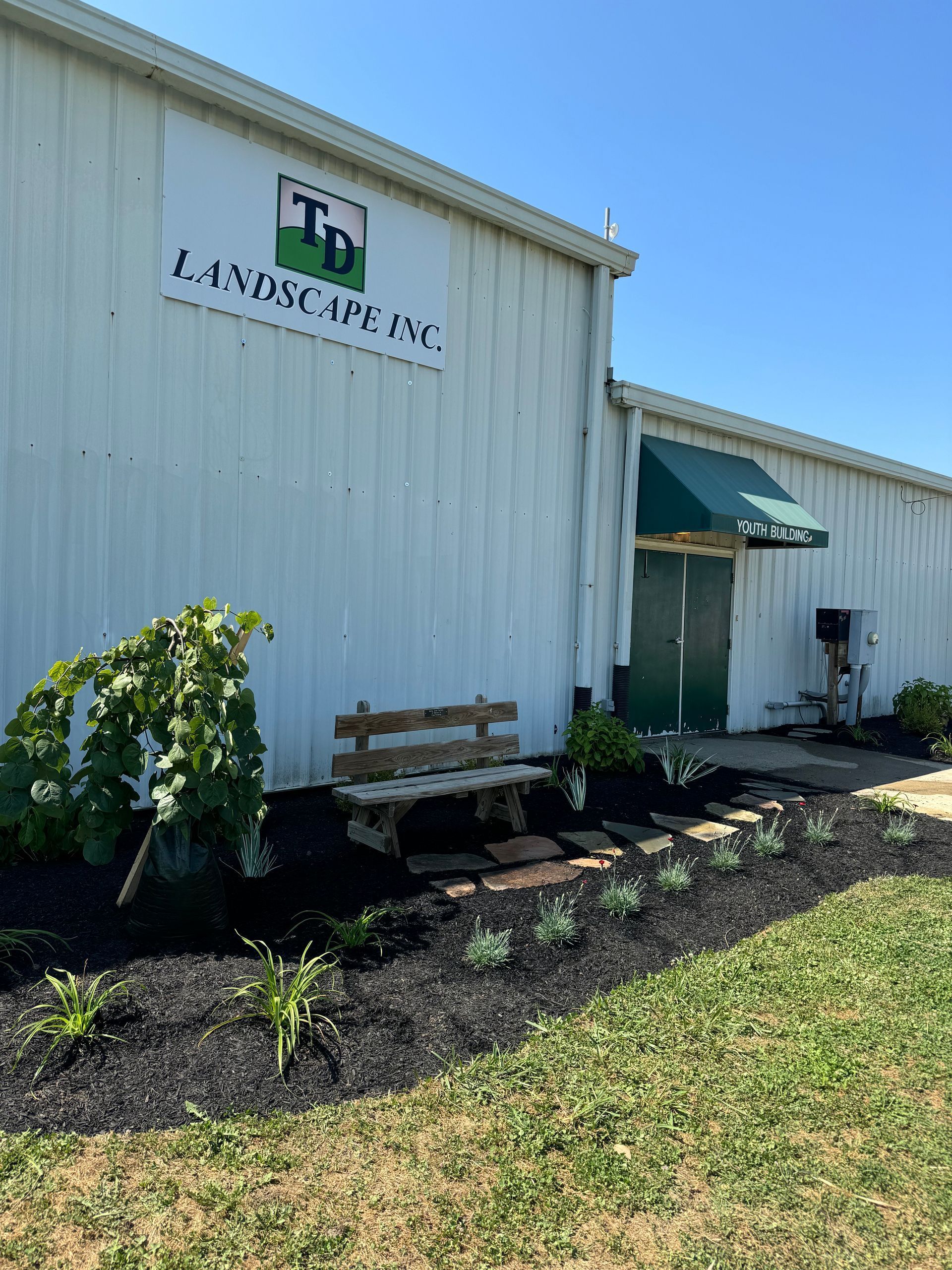White building with a green awning, bench, and garden beds under a clear blue sky.