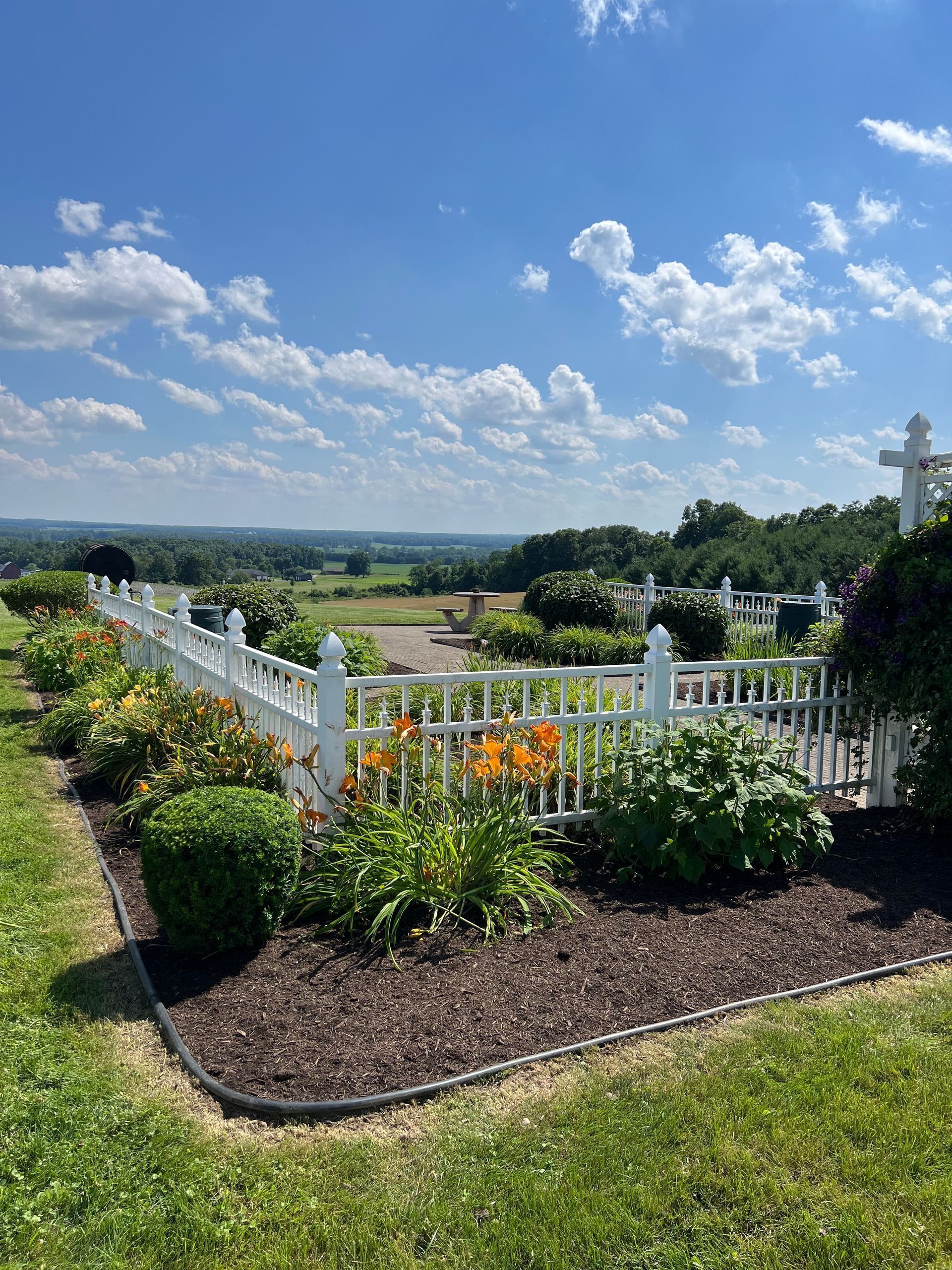 White garden fence with flowers and a cross overlooking a sunny countryside under a blue sky with clouds