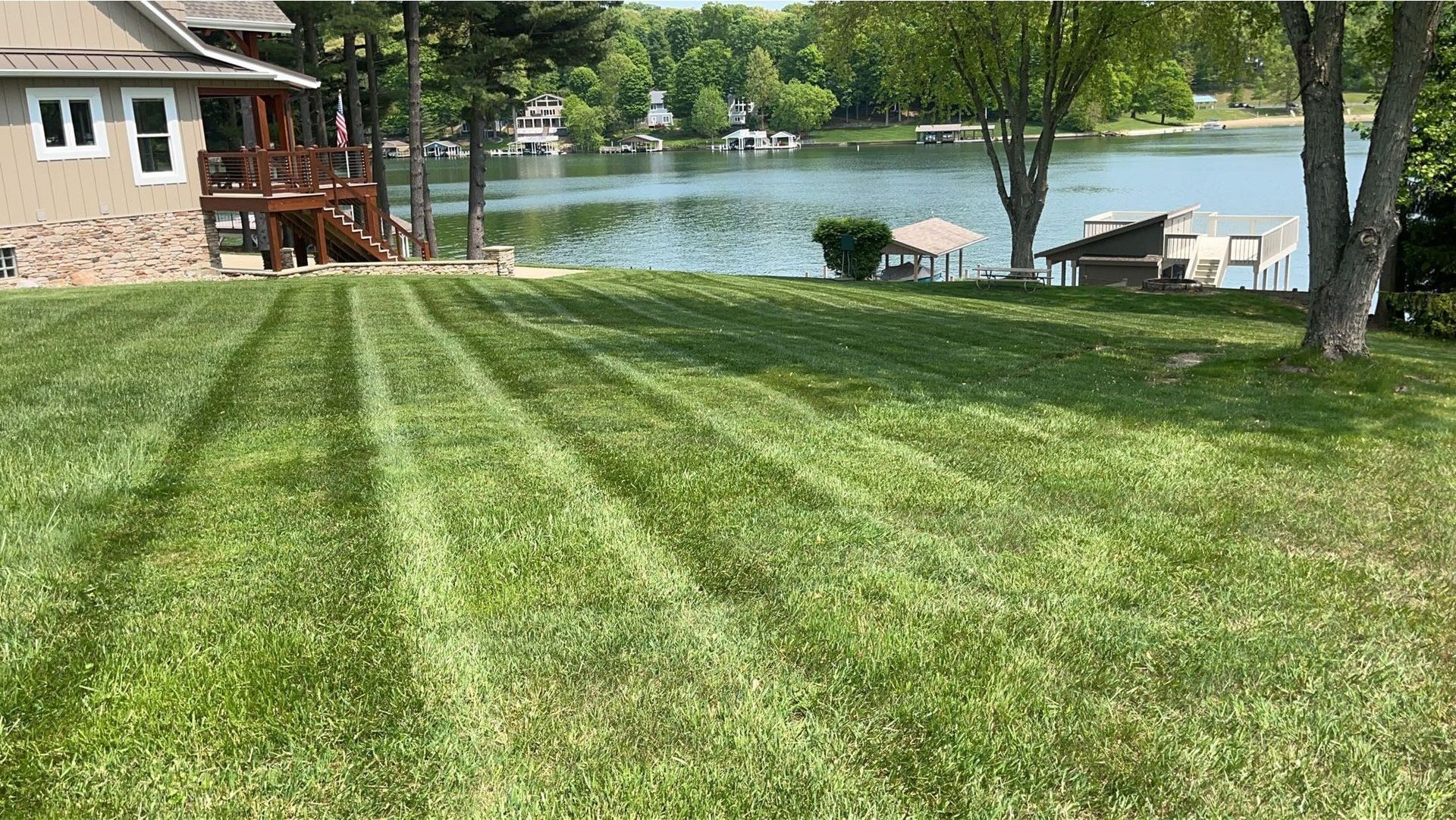 Freshly mowed lawn leading to a lakeside dock, patio, and house with trees nearby