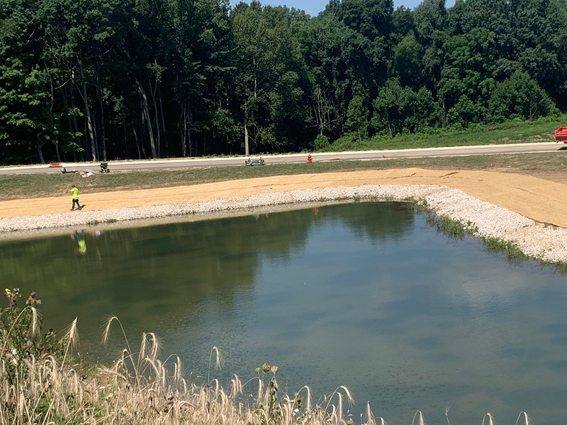 Small pond at a sandy construction site bordered by trees, with a person in a safety vest near the shore