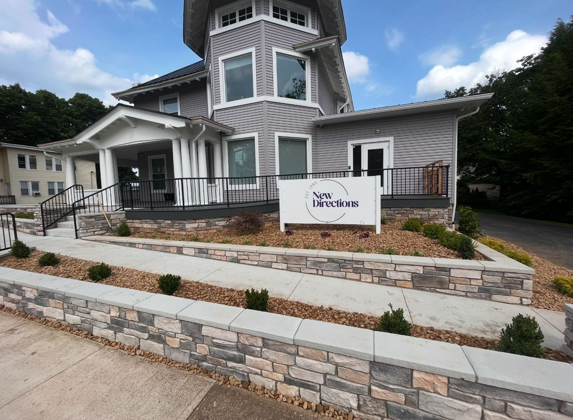 Stone building with wraparound porch, white trim, and a sign in front, set beside a landscaped stone wall.