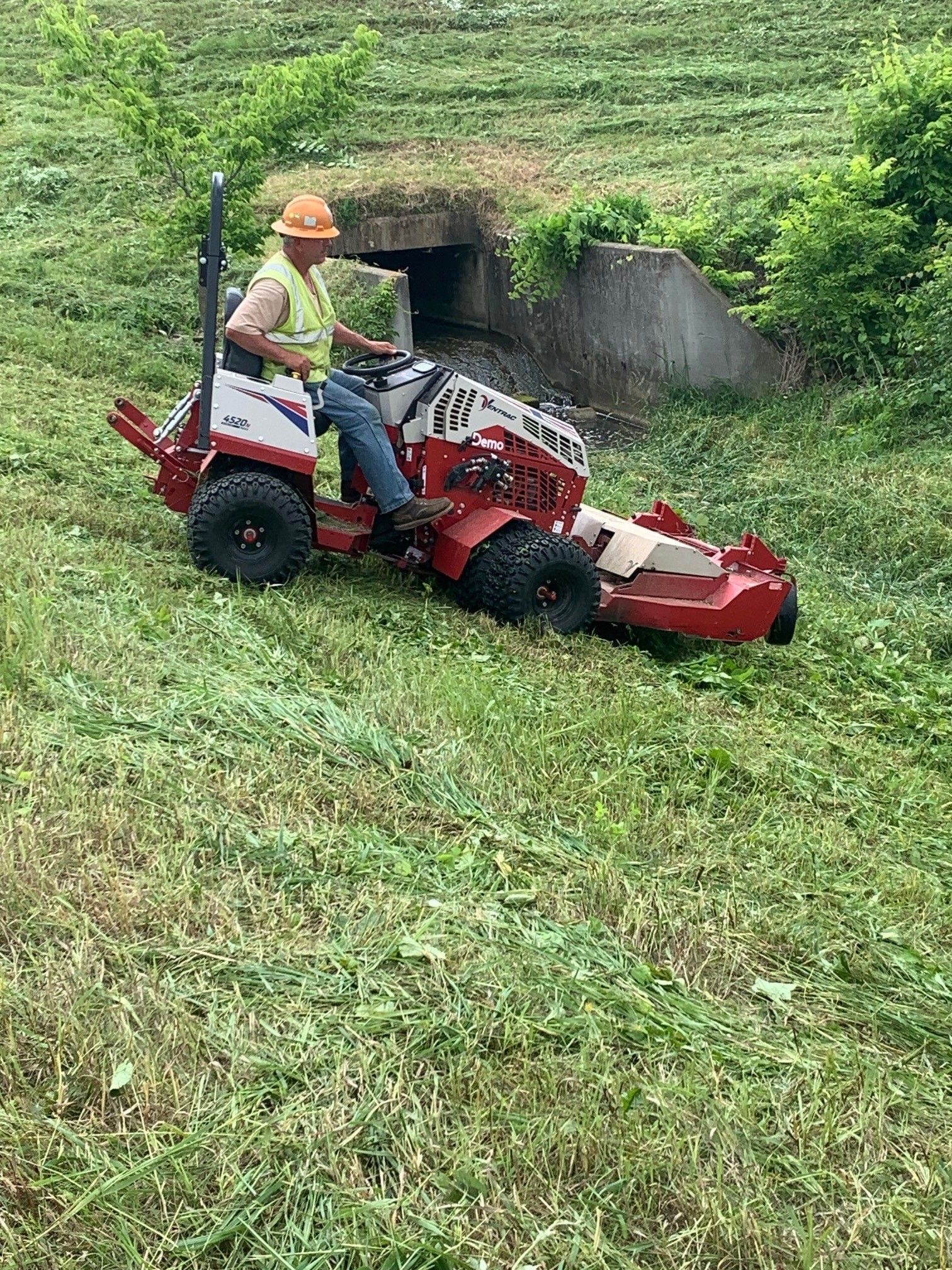 Person mowing a steep grassy slope on a red riding mower near a field edge
