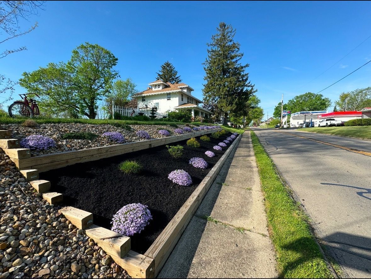 Flower-lined stone wall beside a road, with a white house and tall trees under a clear blue sky.