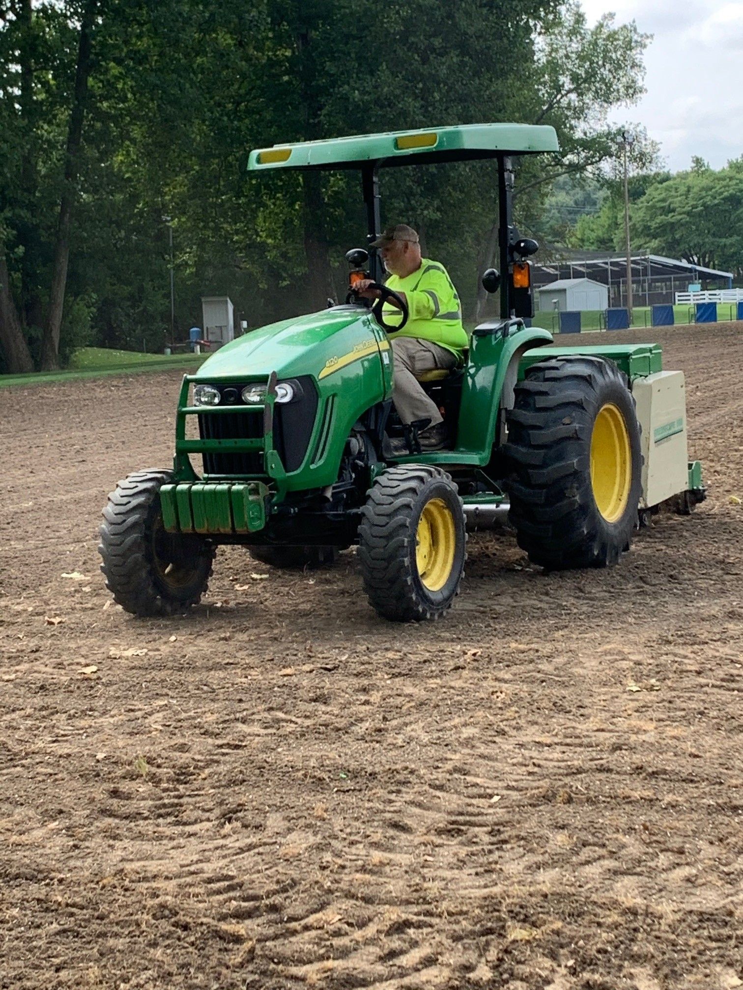 Green tractor with a driver on a dirt field, towing a large rear implement under a canopy.