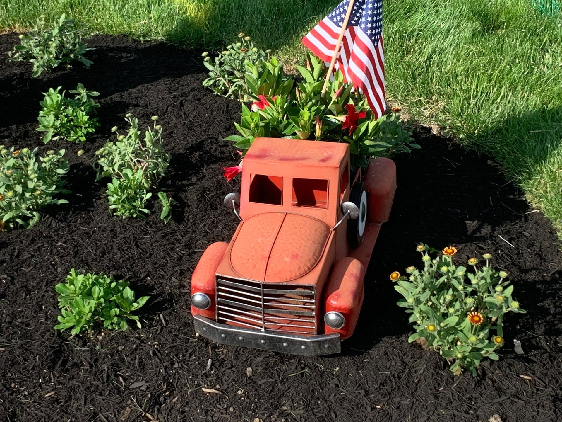 Red toy truck planter in a garden bed with flowers and a small American flag