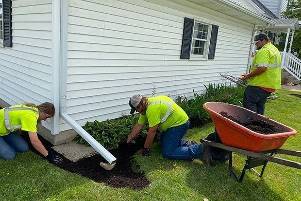 Three workers in neon shirts installing a white downspout beside a house, with a wheelbarrow nearby