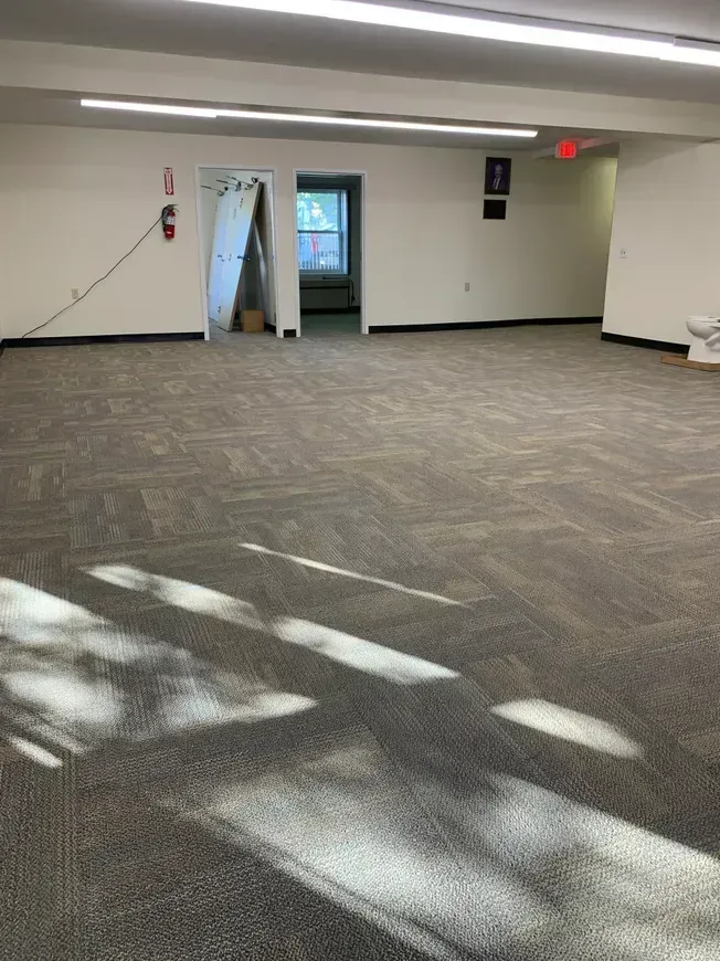 Empty room with neutral-toned carpet, white walls, and bright overhead lighting. Two doorways and a fire extinguisher are visible.