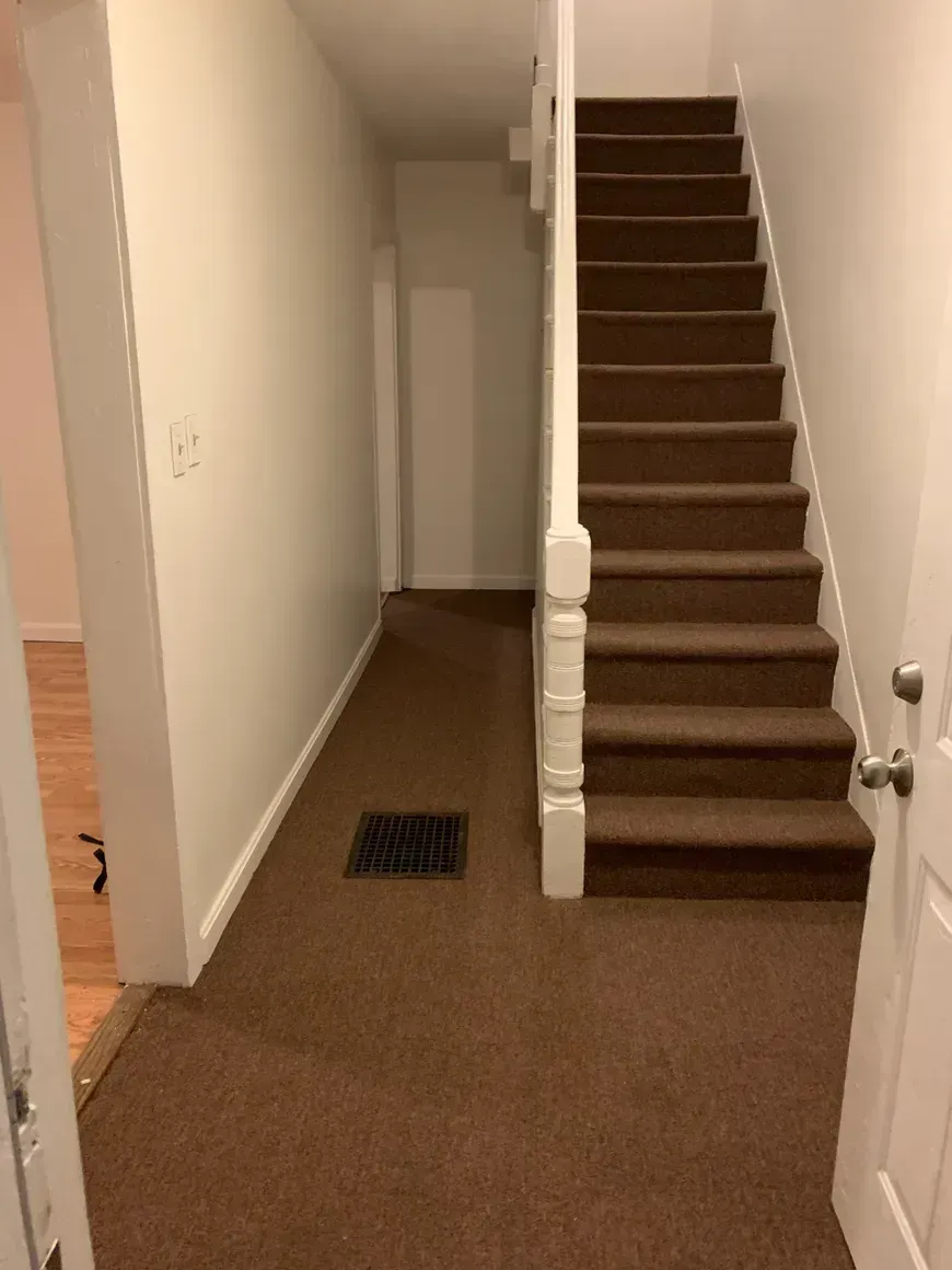 Brown carpeted stairs next to a hallway with beige walls and a doorway on the left.