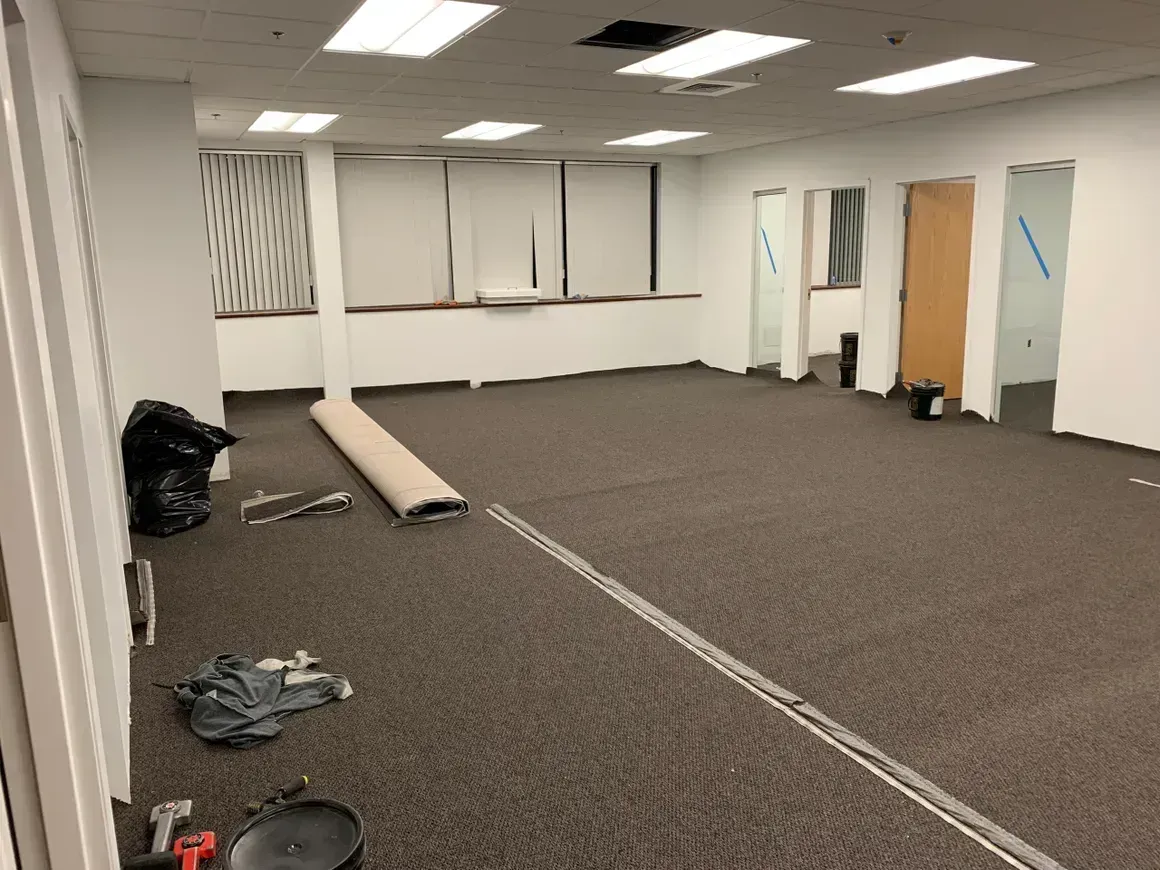 An empty office room with dark carpet, white walls, and a window. Tools and materials are scattered on the floor.