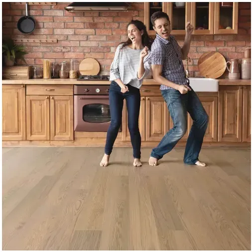 A couple dances barefoot in a kitchen with wood floors and brick wall, joyfully moving their arms.