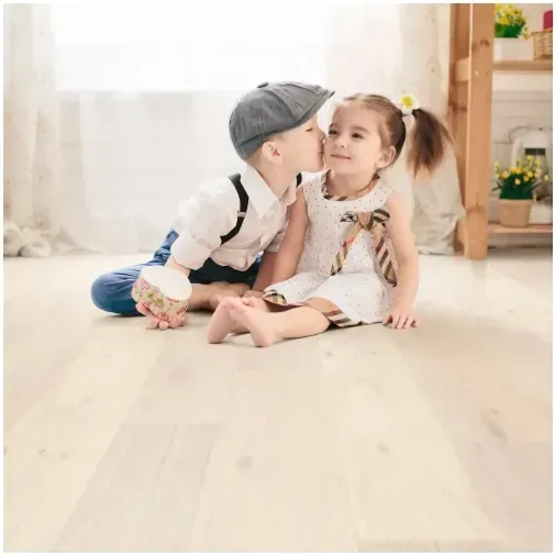 Boy in newsboy cap kisses girl on cheek, both sit on a light wood floor.