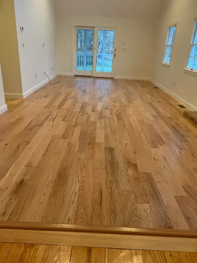Light wood flooring in a mostly empty room with white walls, two windows, and a glass door.