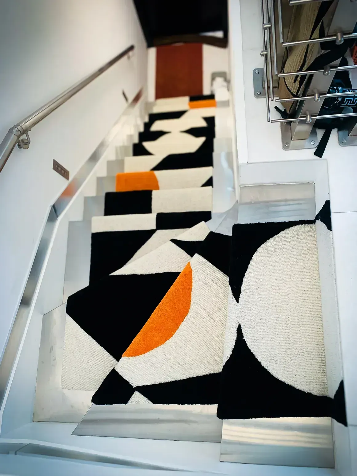 Staircase with geometric patterned carpet in black, white, and orange. White walls and metal handrail.