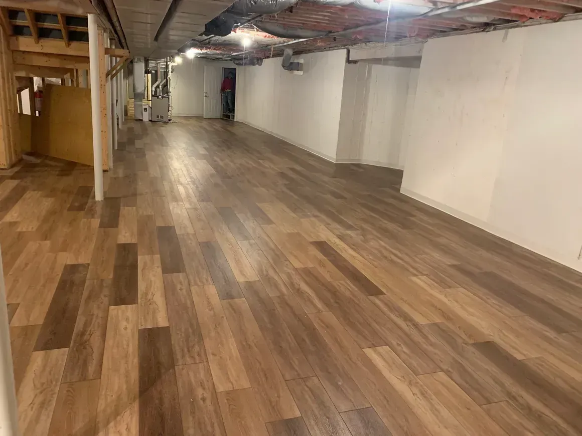 Spacious basement room with wood-look flooring, white walls, and exposed ceiling infrastructure.