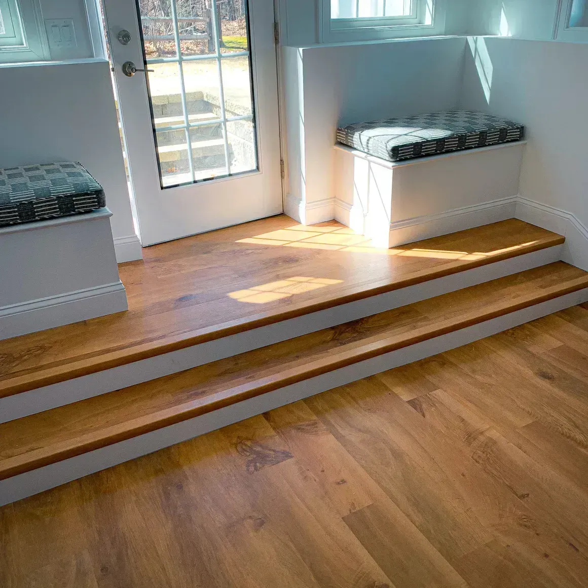 Wooden steps leading to a door with built-in seating on either side. Sunlight streams through the door.