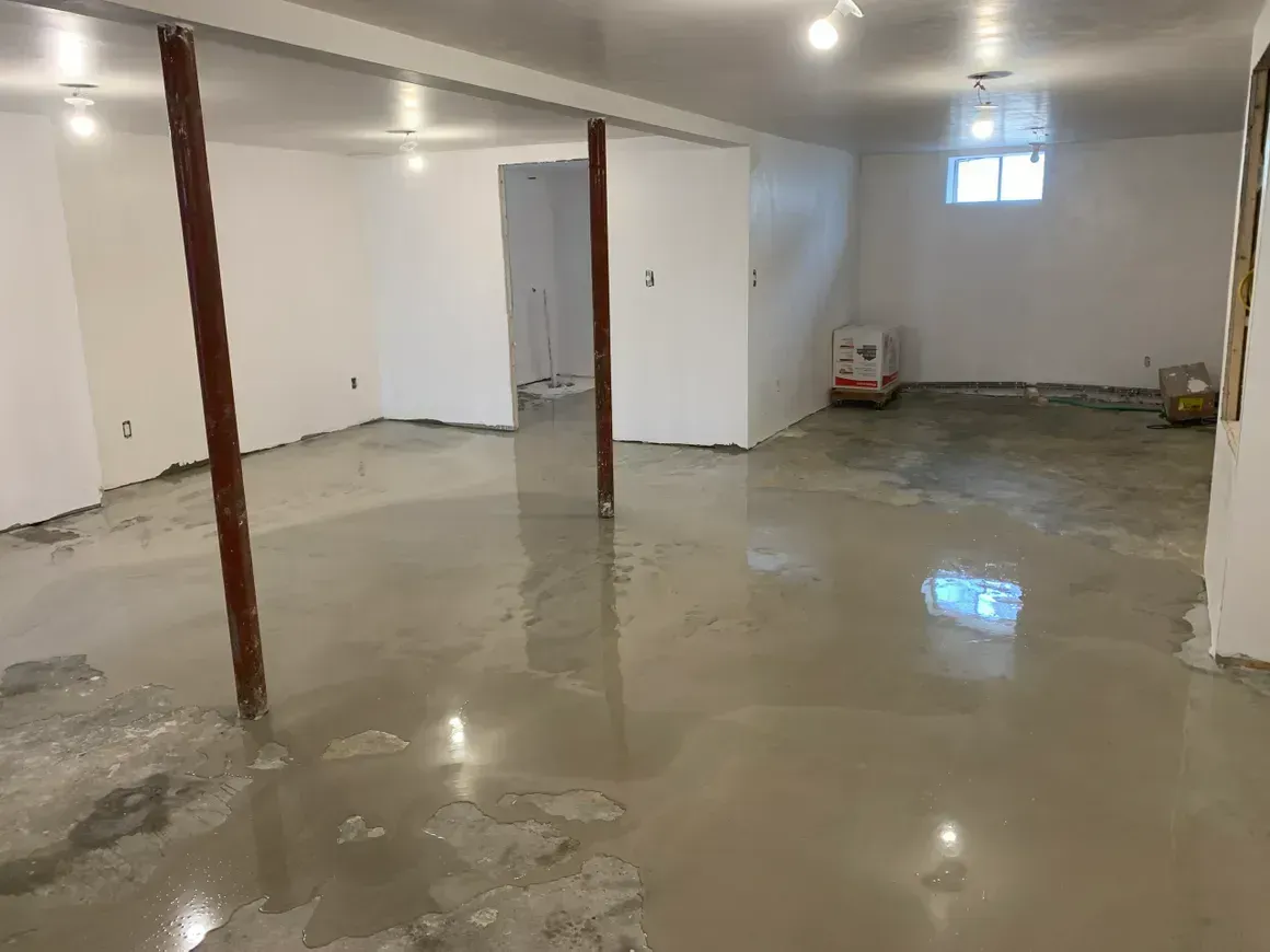 Basement with wet, smooth concrete floor; support beams; white walls; and overhead lights.